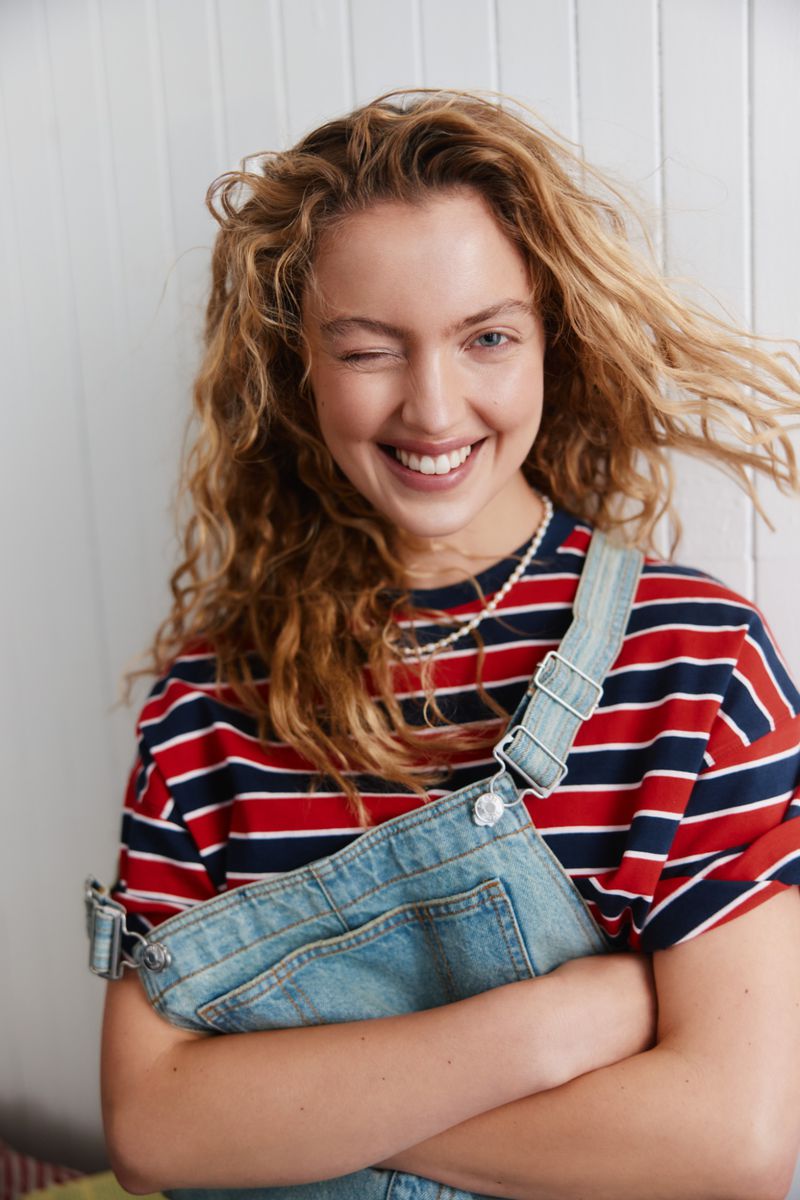 Young woman with curly hair wearing striped shirt and overalls winking at the camera in an indoor setting with white wall backdrop.