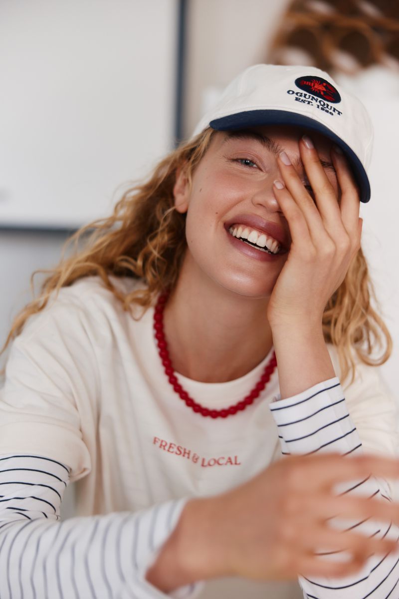 Smiling woman in cap poses indoors, wearing a white t-shirt and red necklace with white walls visible in the background.