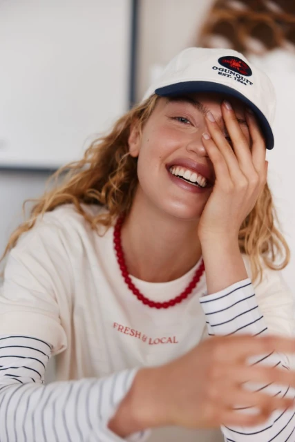 Smiling woman in cap poses indoors, wearing a white t-shirt and red necklace with white walls visible in the background.