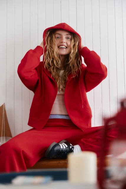 Smiling woman in a red hooded jacket and matching pants sits indoors, showcasing a casual fashion style with wavy blonde hair.