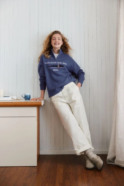 A young woman with curly hair leans against a white wall wearing a blue sweater and white pants, showcasing a casual indoor fashion style.