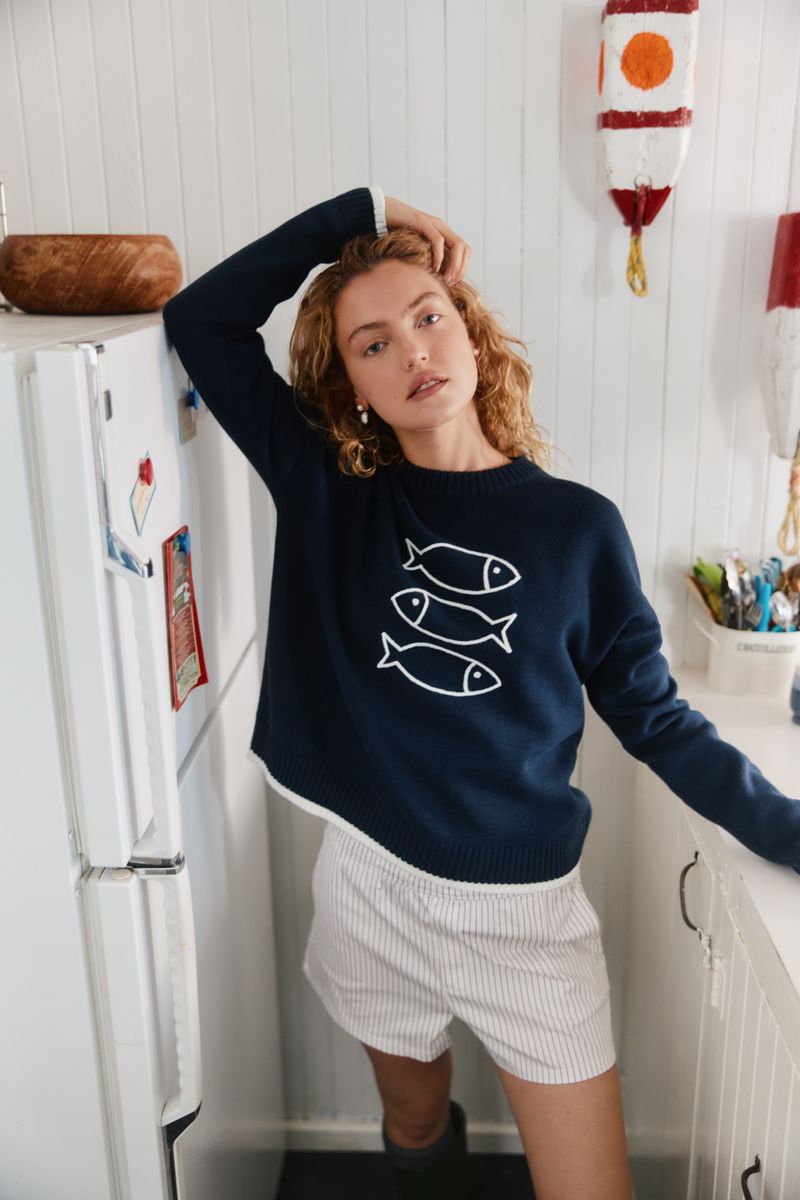 Blonde model in a navy sweater with a fish design poses indoors in a white kitchen with an off-white wall and nautical decor.