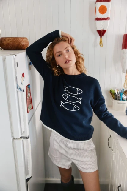 Blonde model in a navy sweater with a fish design poses indoors in a white kitchen with an off-white wall and nautical decor.