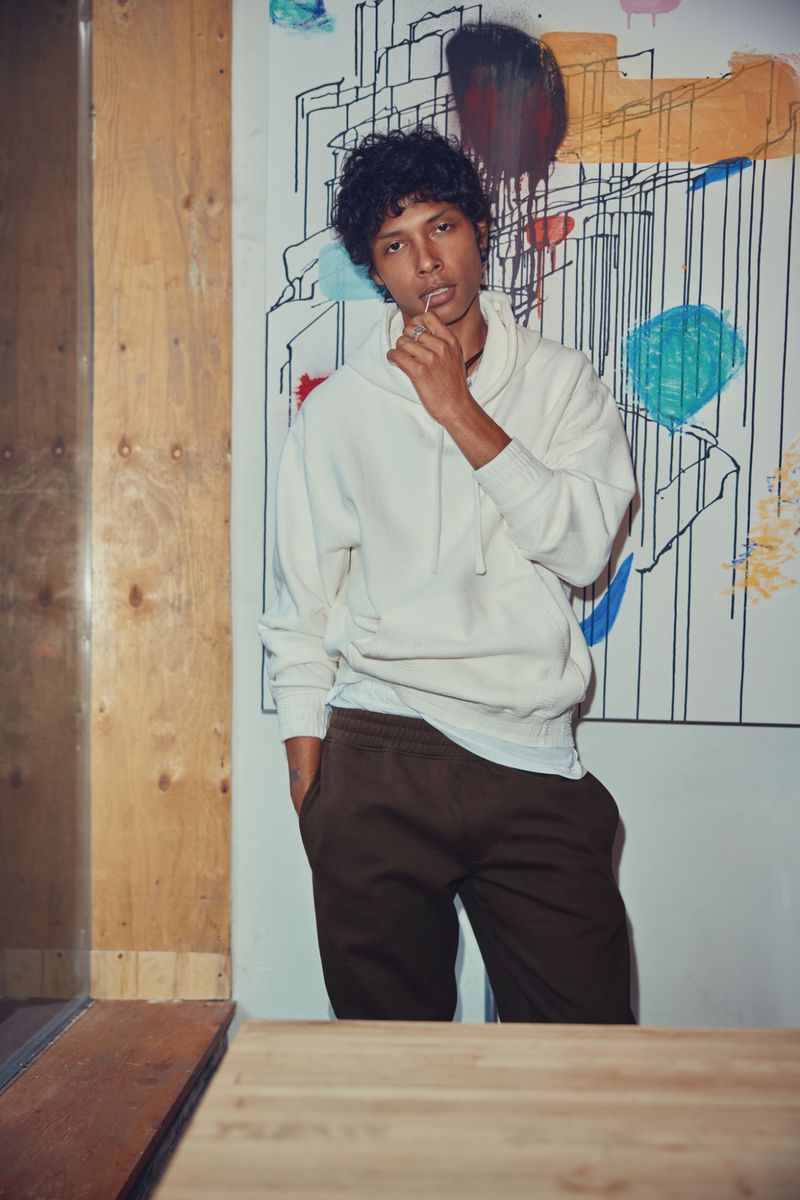 Young adult male posing in white hoodie and brown pants, standing in an indoor setting with an abstract art background.