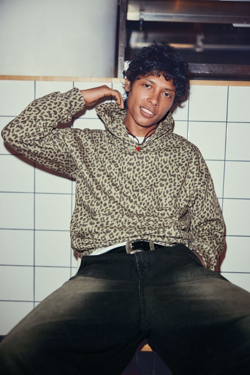 Young man with curly dark hair, wearing a leopard print hoodie and jeans, smiles in an indoor setting with white tiled walls.