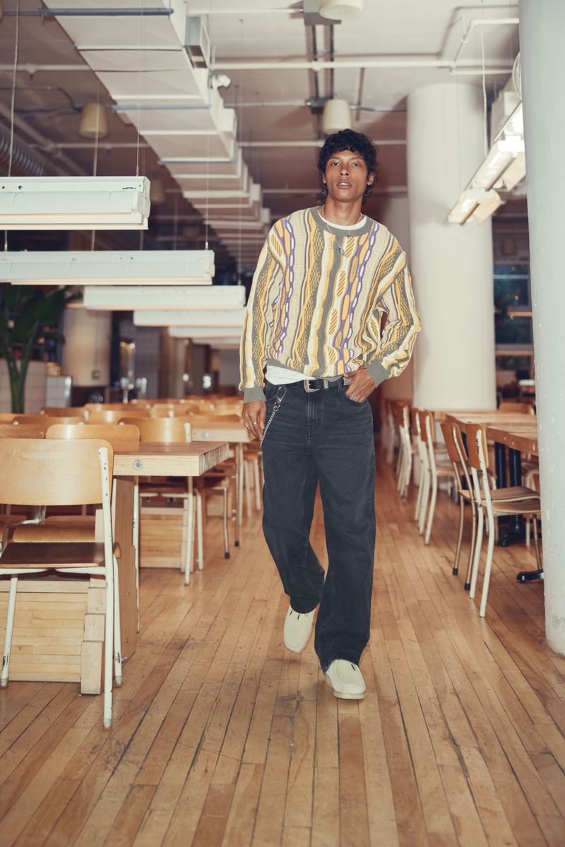 Young man in patterned sweater and dark pants walking in a brightly lit building with rows of tables and chairs.