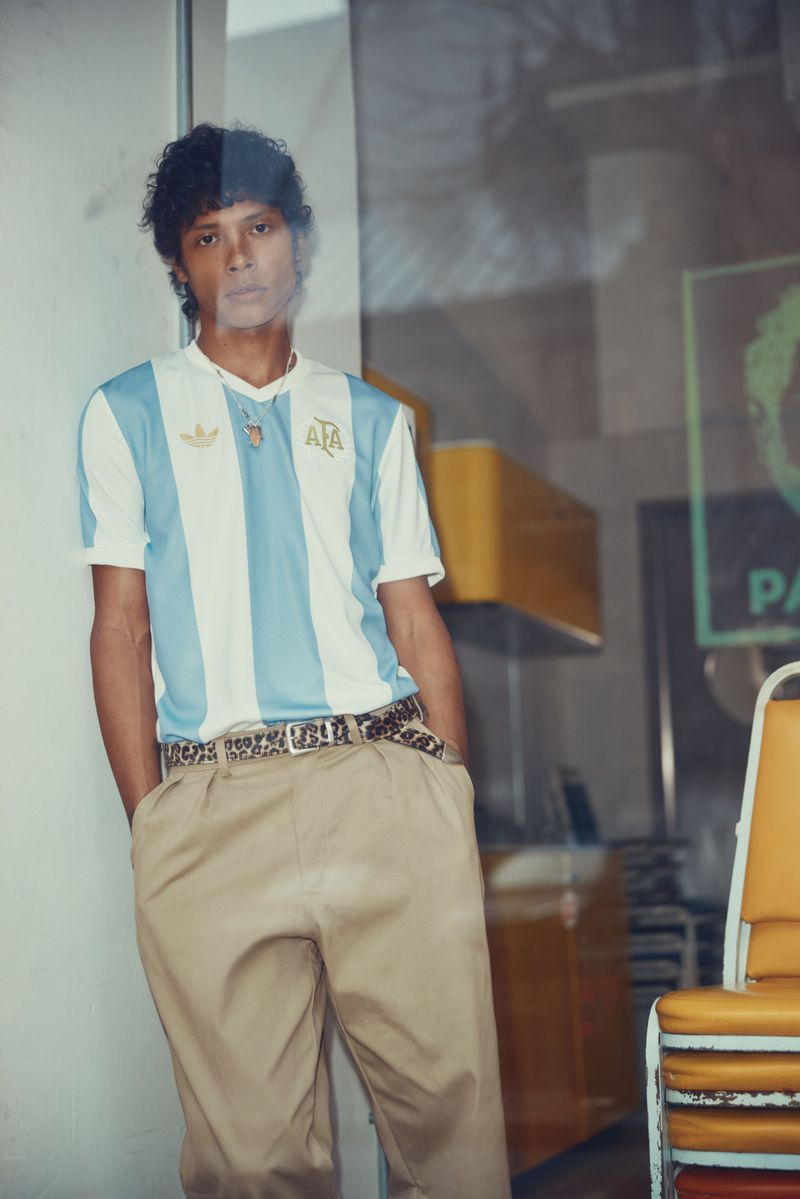 Man posing in an Argentina soccer jersey, khaki pants and patterned belt, standing against a wall in an indoor setting with yellow chairs.