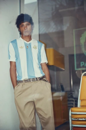 Man posing in an Argentina soccer jersey, khaki pants and patterned belt, standing against a wall in an indoor setting with yellow chairs.