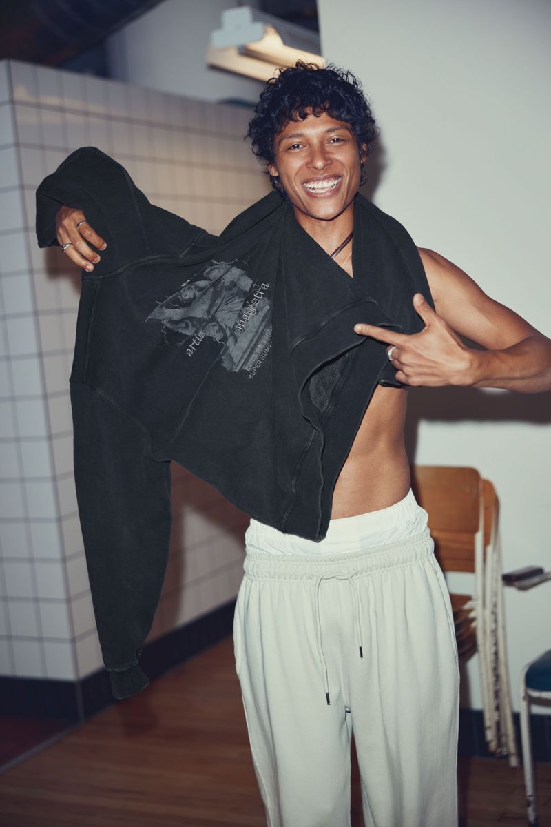 Smiling adult male pointing at a graphic printed dark hoodie in an indoor environment with a chair and white tiled walls.