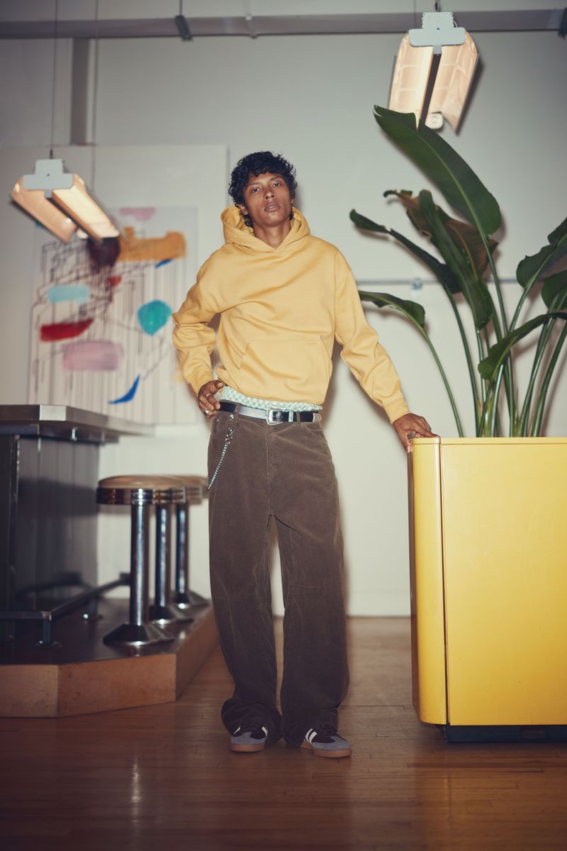 Full-length shot of a young adult wearing a yellow hooded sweatshirt, brown corduroy pants, and sneakers in an indoor setting with plants.