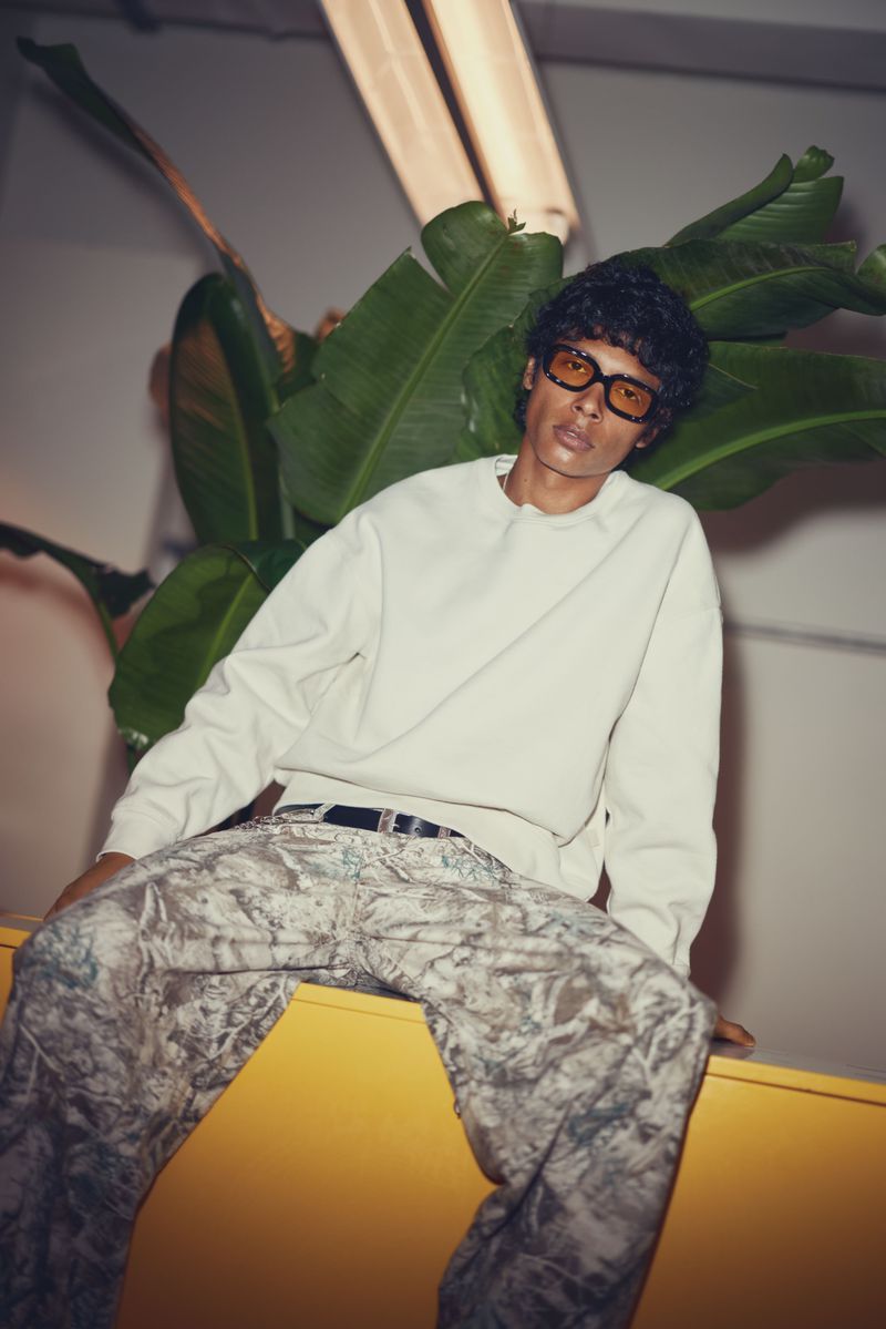Young man in a light sweatshirt and camouflage pants sits on a yellow surface indoors with indoor lighting and a plant in the background.