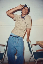 Young male model wearing a beige t-shirt, blue jeans, and cap leaning against a chair for Simons Canada DJAB '24.