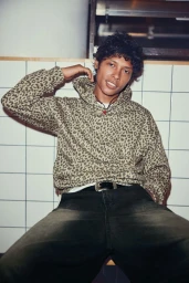 Young man wearing a leopard print hoodie and dark pants posed against a white tiled wall