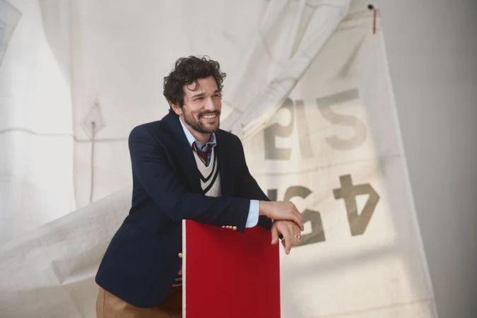 A smiling man in a blazer and sweater vest leans against a red chair against a white sailboat sail backdrop indoors.