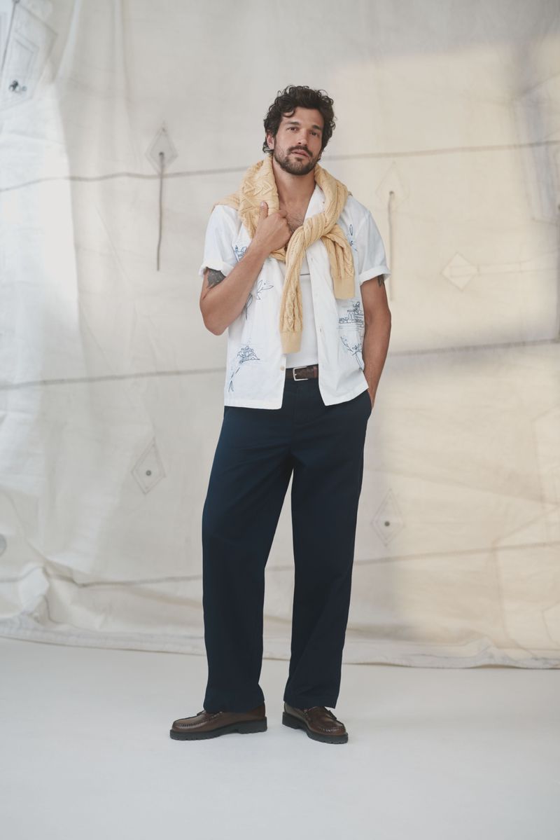 Man in casual outfit stands in a relaxed pose. He wears a white shirt, blue pants, brown shoes, and a yellow sweater draped over his shoulders.