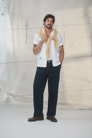 Man in casual outfit stands in a relaxed pose. He wears a white shirt, blue pants, brown shoes, and a yellow sweater draped over his shoulders.