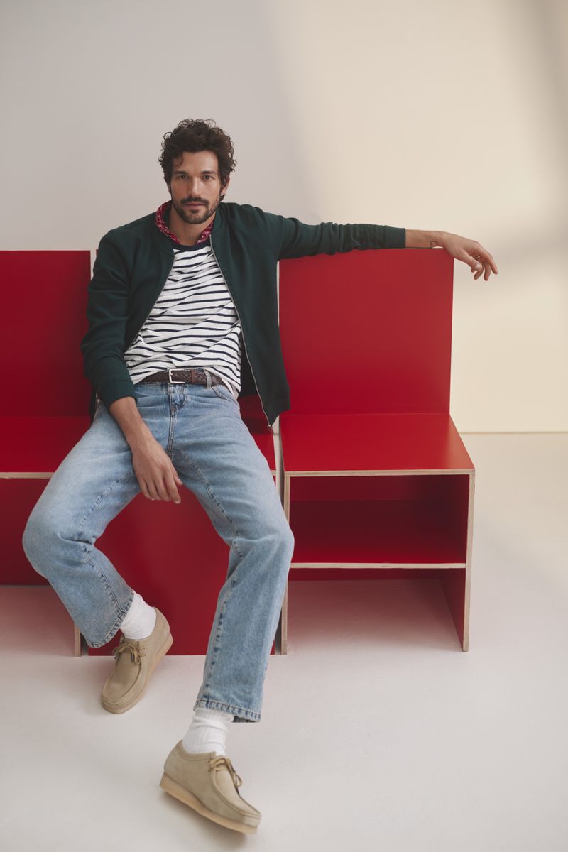 A stylish adult male sits on a red bench indoors wearing a green jacket, striped t-shirt, jeans, and shoes in a casual pose.