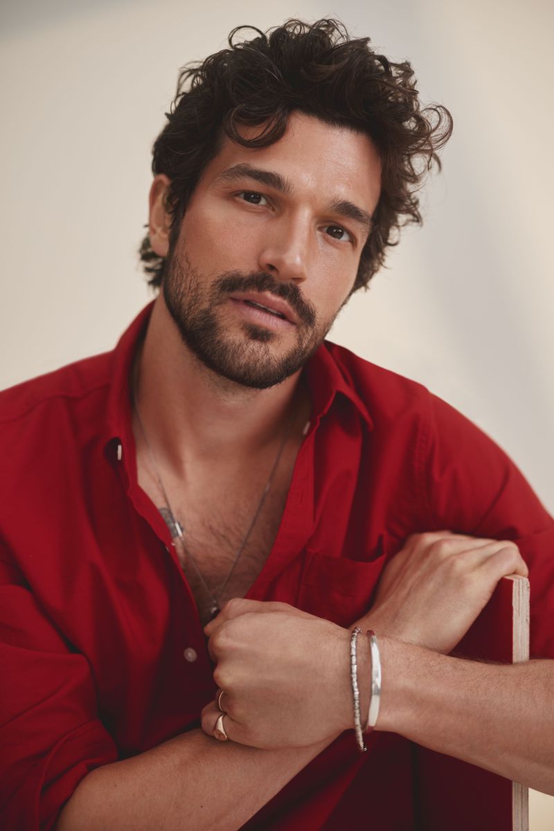 Portrait of a man with dark curly hair wearing a red shirt and silver bracelets against a plain light background