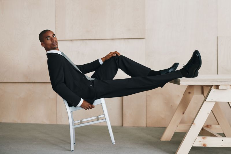A dark-skinned man in a black suit sits in a white chair with his legs propped up on a wooden table, posing in an indoor setting.