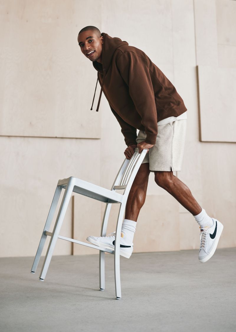 An adult Black male is posing indoors, leaning on a white chair. He's wearing a brown hoodie, light-colored shorts, and white sneakers.