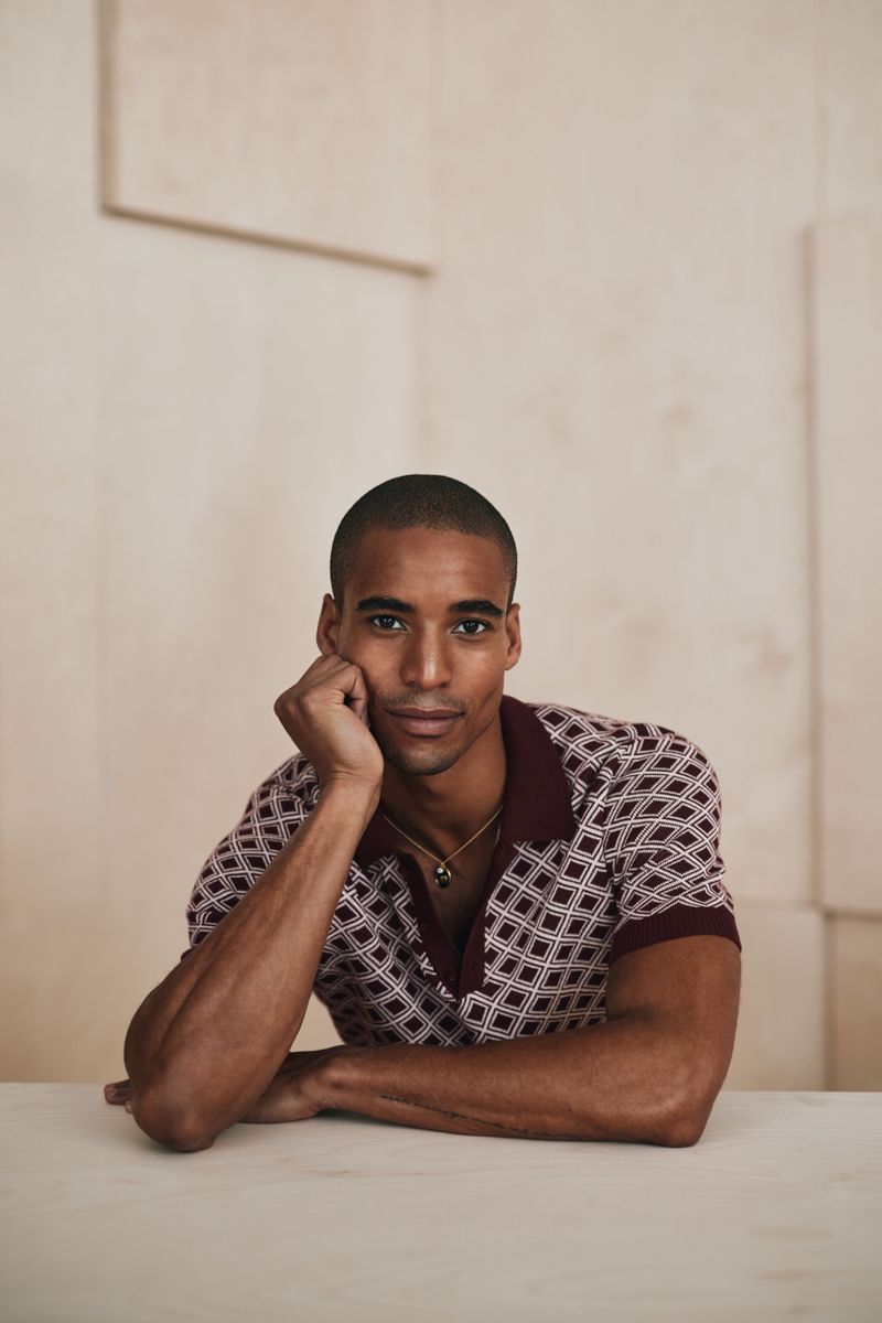 Portrait of a handsome man with dark hair and a patterned polo shirt, resting his head on his hand at a light-colored table indoors.