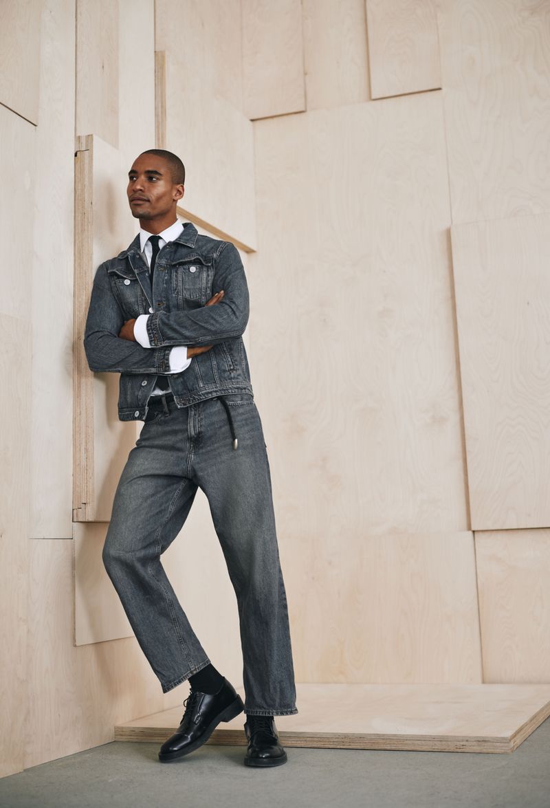 An adult male poses indoors wearing a grey denim jacket and matching jeans, a white dress shirt, black necktie, black dress shoes, and black socks.