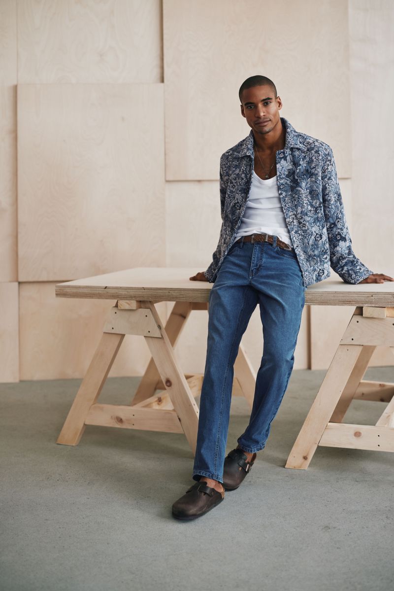 A stylish adult male is posing indoors, wearing a white tank top, blue jeans, a patterned shirt-jacket, and brown clogs, leaning on a wooden table.