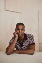 Model Malik Lindo wearing a patterned burgundy polo shirt resting his chin on his hand against a neutral studio backdrop for Simons Canada.