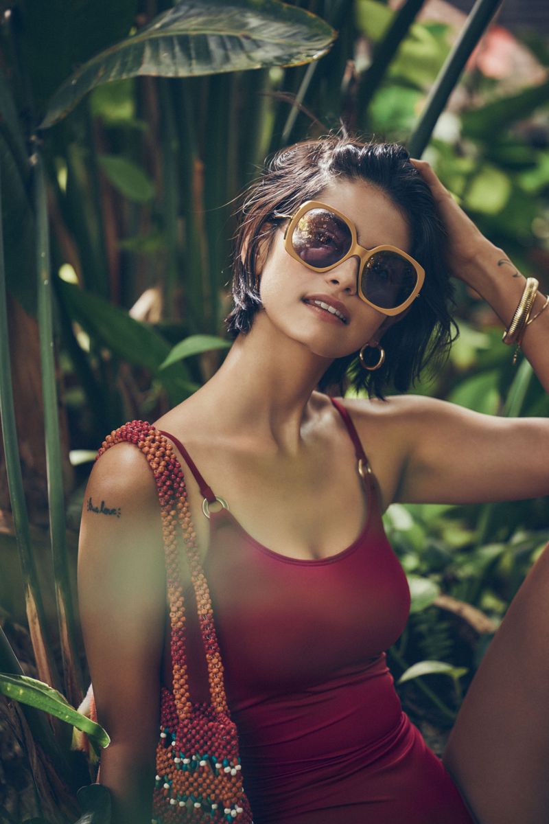 Woman in a red swimsuit and sunglasses posing among tropical foliage. She is carrying a beaded purse, looking towards the camera.