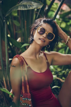 Woman in a red swimsuit and sunglasses posing among tropical foliage. She is carrying a beaded purse, looking towards the camera.