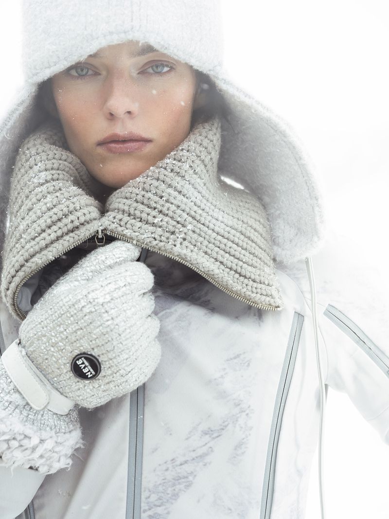 A woman in a white winter hat, textured knit scarf, and matching gloves, wearing a white winter jacket in a brightly lit outdoor setting.