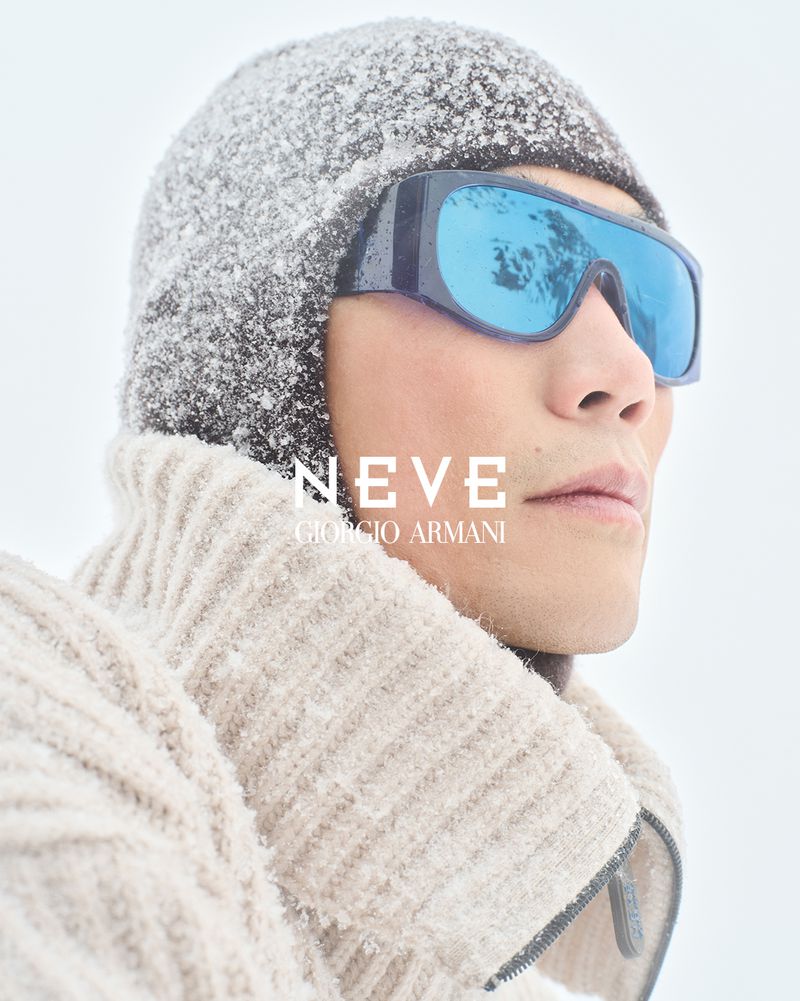 Portrait of an adult male wearing sunglasses, knit cap, and wool sweater, with a snowy white outdoor background, and 'NEVE GIORGIO ARMANI' text.