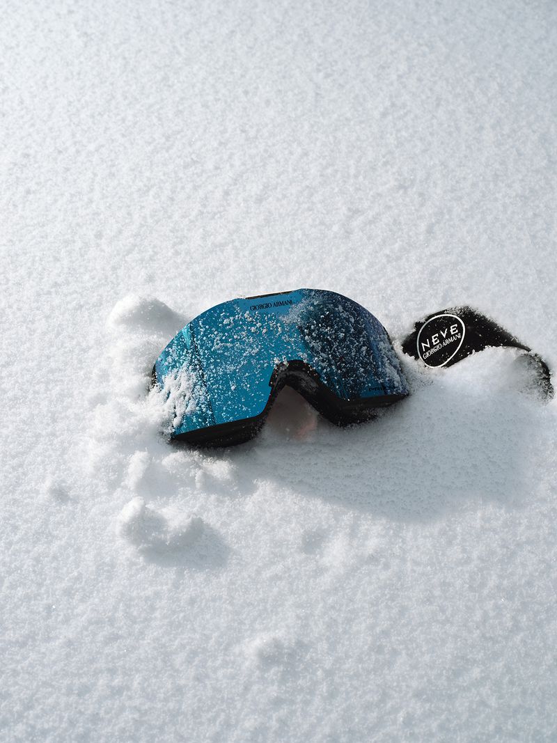 Close-up of blue-tinted ski goggles dusted with snow, resting on a snowy surface with a visible brand tag, outdoors.