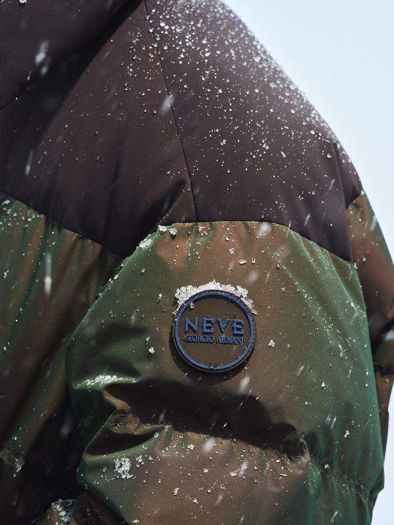 Close-up of a green and brown puffer jacket with a Neve Giorgio Armani logo in falling snow against a bright, neutral backdrop.