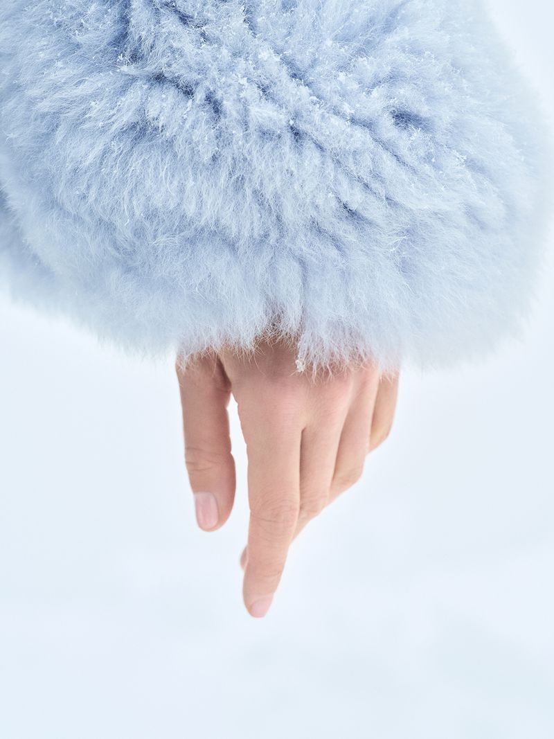 Close-up on a woman's hand with pale skin pointing downward, accented by a light blue furry coat sleeve in a soft, bright setting.