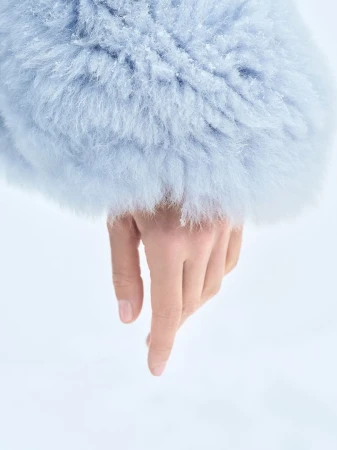 Close-up on a woman's hand with pale skin pointing downward, accented by a light blue furry coat sleeve in a soft, bright setting.