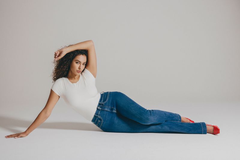 A curly-haired woman in a white t-shirt and blue jeans poses while lying on her side against a light-colored studio backdrop.