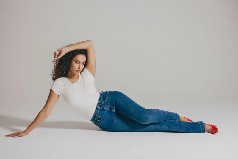 A curly-haired woman in a white t-shirt and blue jeans poses while lying on her side against a light-colored studio backdrop.