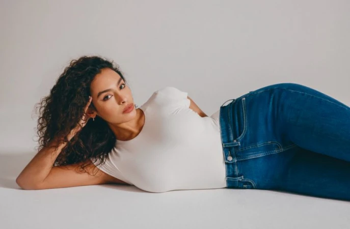 Model Kenza Safsaf reclining in white t-shirt and blue denim jeans against a seamless white studio background.