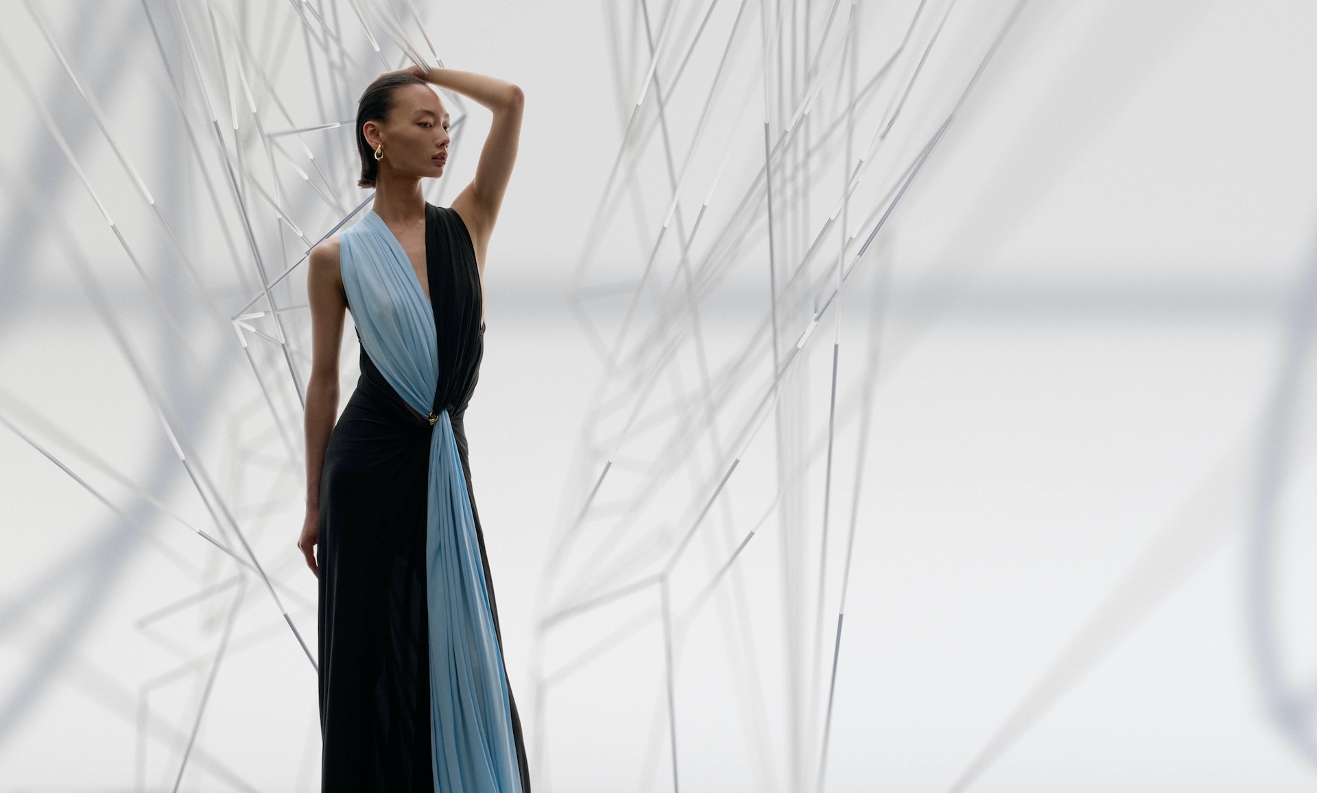 Elegant woman poses in a black dress with a blue drape, set against a backdrop of geometric white rods, fashion photography.