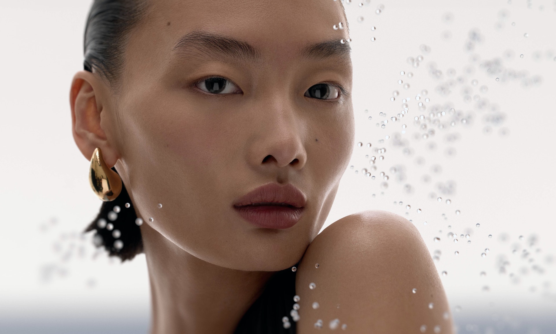 Close-up portrait of an Asian adult female model with gold earring, clean skin, and water droplets, shot in a bright, neutral studio.