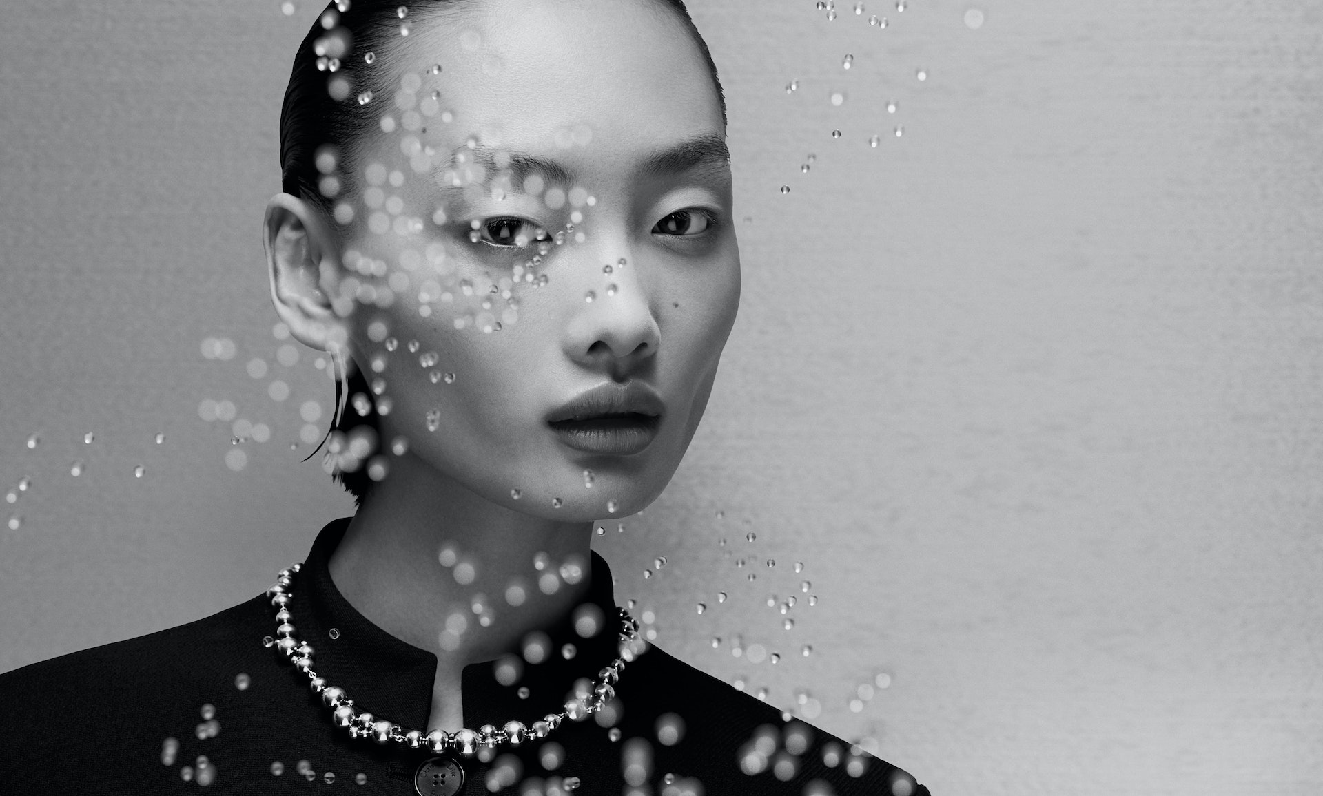 Monochrome portrait of an Asian woman with water droplets, wearing a black collared shirt and necklace against a light gray background.
