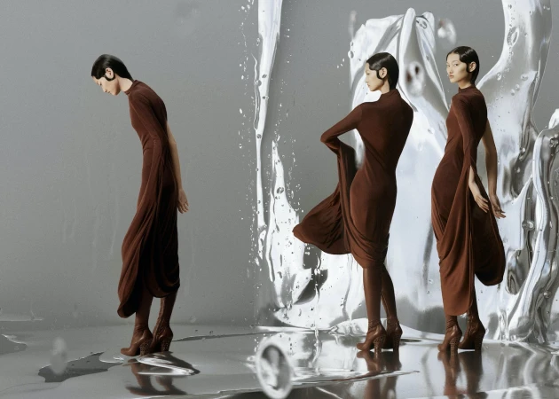 Three women pose in brown dresses with sculptural silver backdrop. Fashion shoot with reflective surface and elegant, contemporary design.