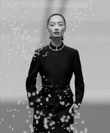 Model in a black buttoned jacket with a pearl necklace against a neutral backdrop, framed by floating water droplets.
