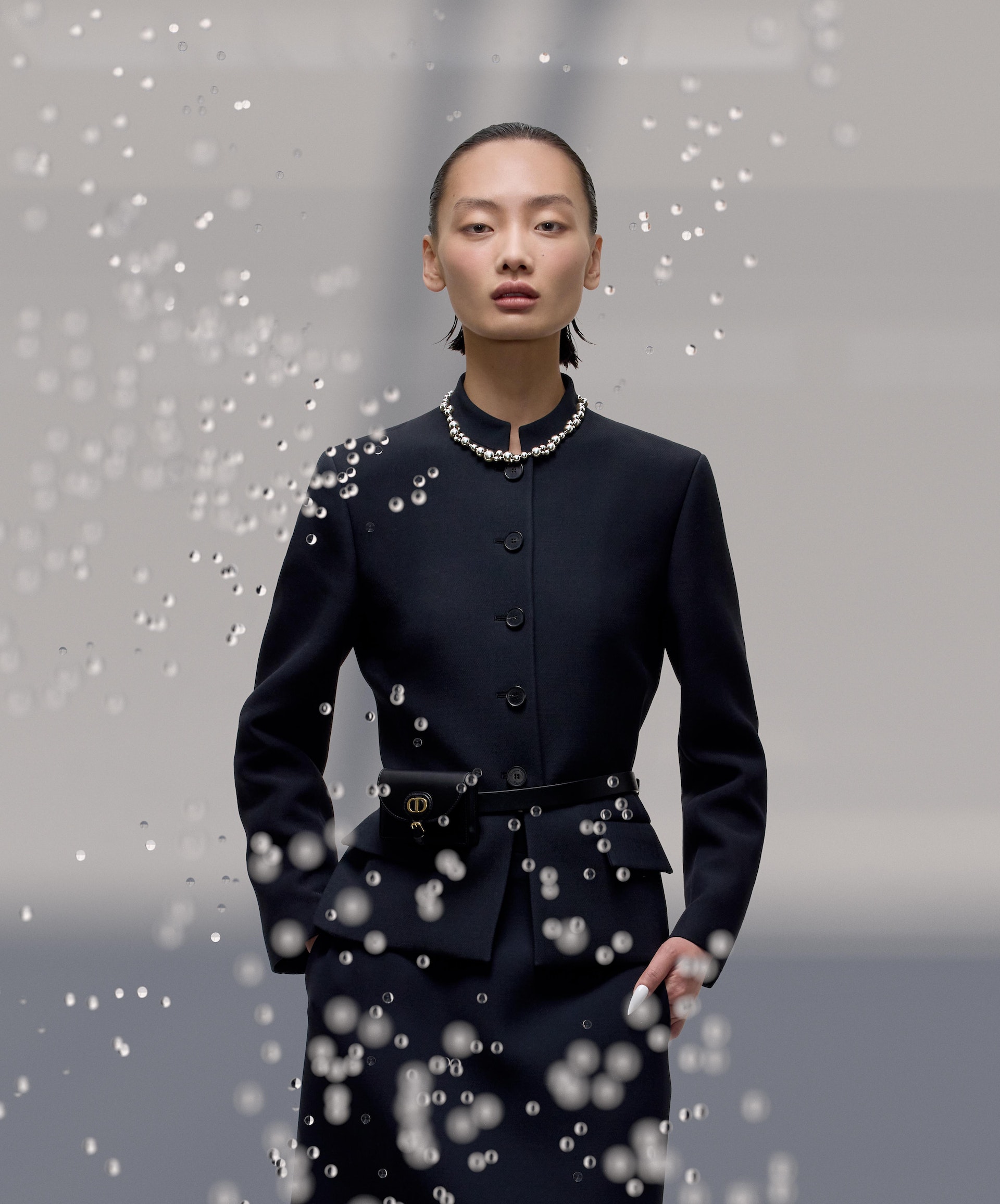 Elegant Asian woman in a navy suit and pearls, adorned with glistening water droplets in a fashion shot with a grey gradient background.