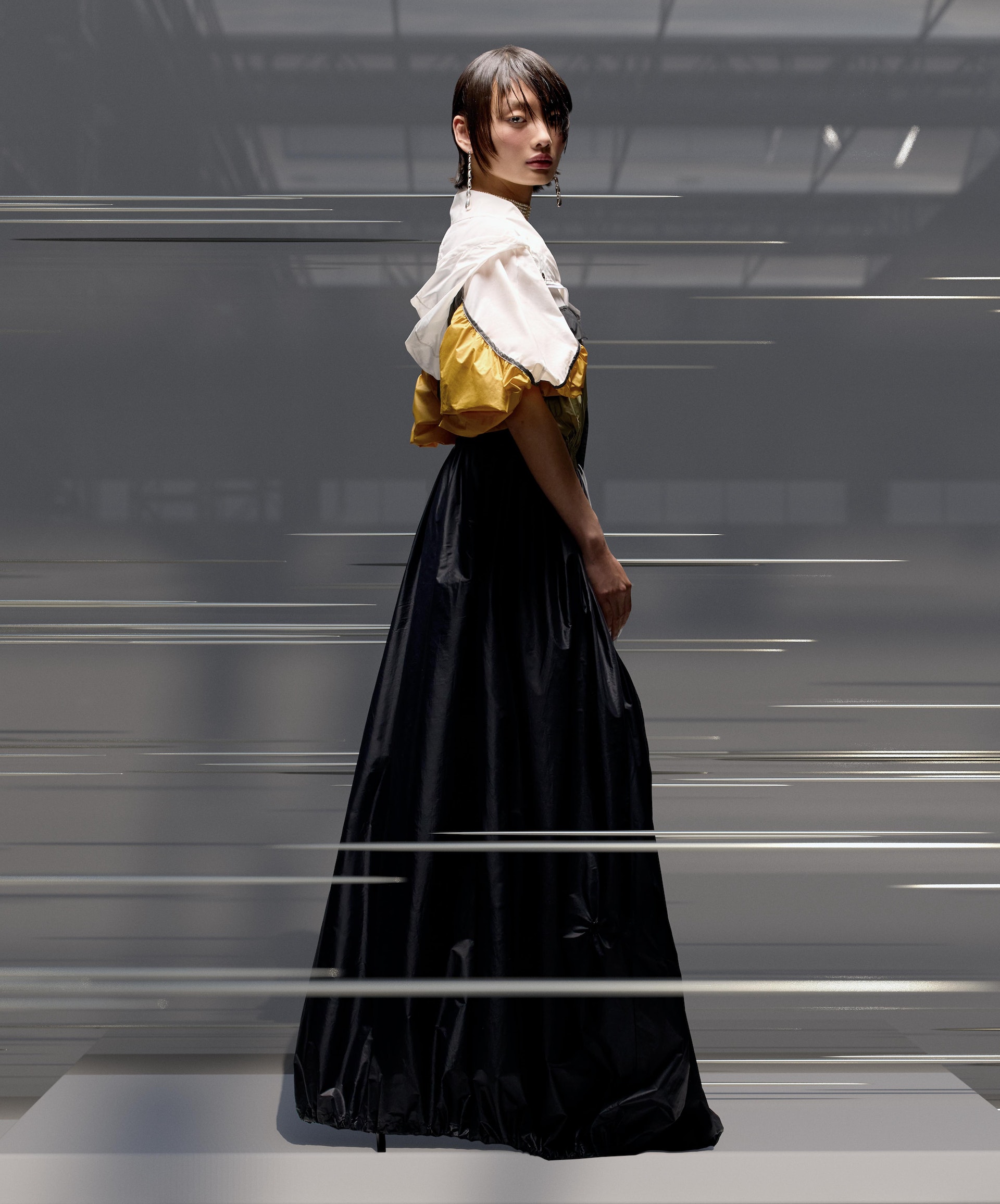 Woman in a white and yellow top with black skirt posing in a studio, looking over her shoulder in a three-quarters view.