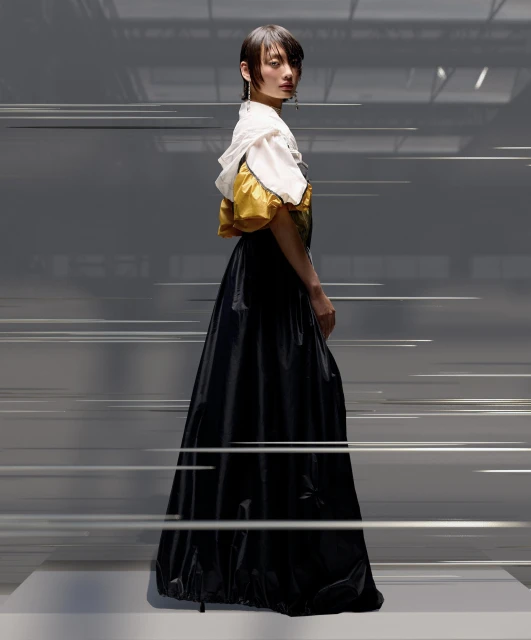 Woman in a white and yellow top with black skirt posing in a studio, looking over her shoulder in a three-quarters view.