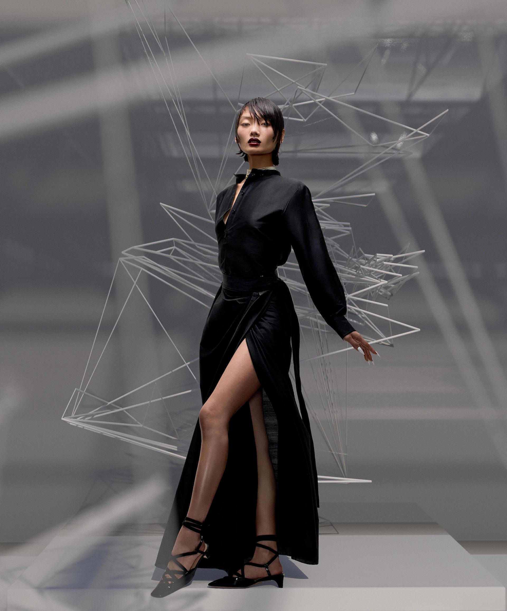 Elegant Asian model poses in a black dress with a high slit, set against a backdrop of a geometric wire sculpture in a studio.