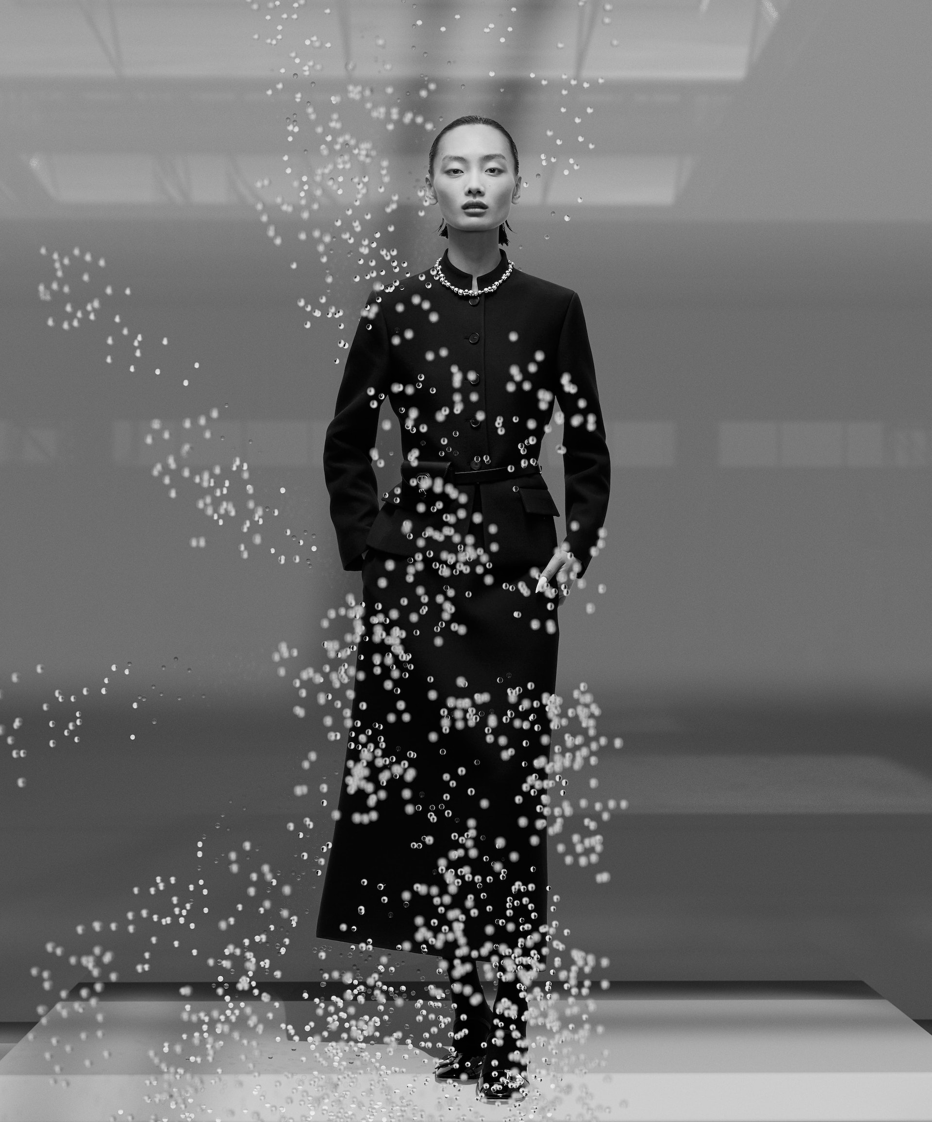 A sophisticated black-and-white studio portrait of an Asian female fashion model wearing a dark belted coat and pearl necklace.