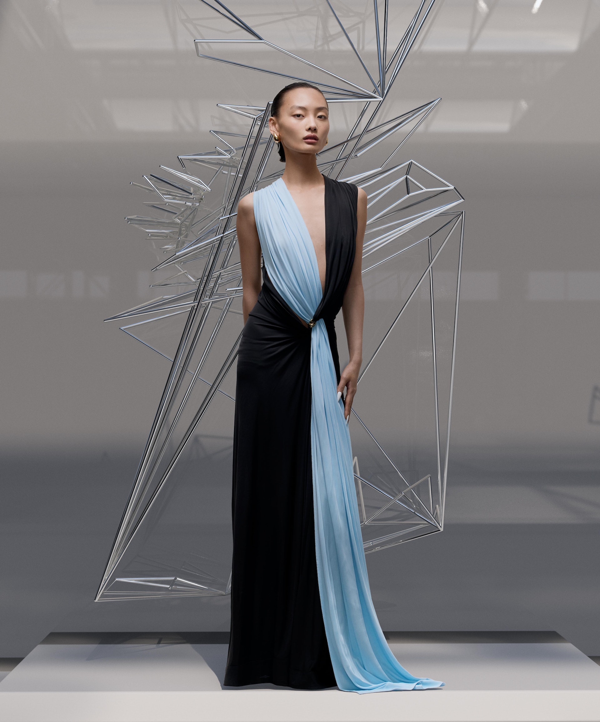 Elegant Asian woman poses in a striking black and blue gown next to modern geometric art installation in a bright, minimalist setting.